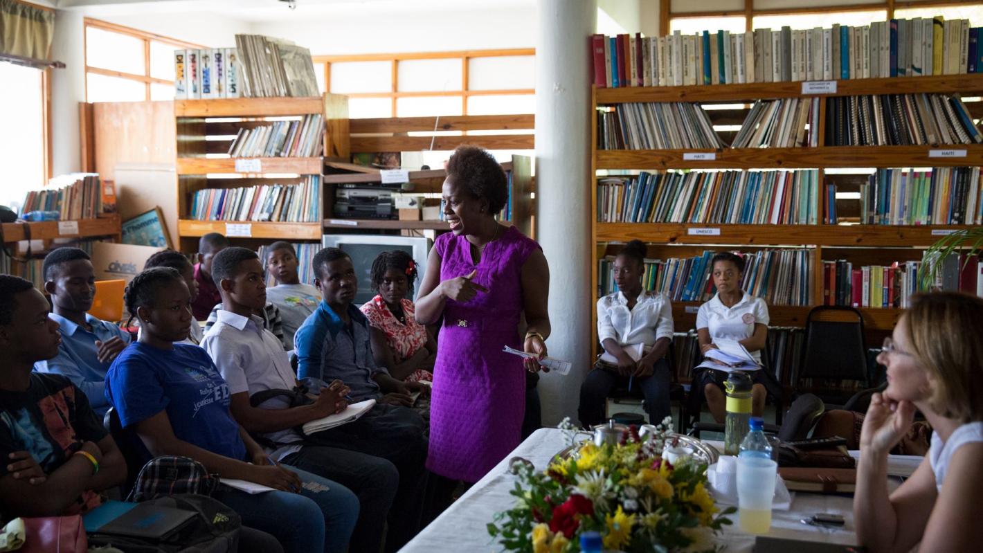 A group of people sitting in a library listening to a woman in a purple dress speaking to the group.
