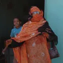 A woman standing in traditional dress holding her arm out. A child in a blue t-shirt is behind the woman's arm.
