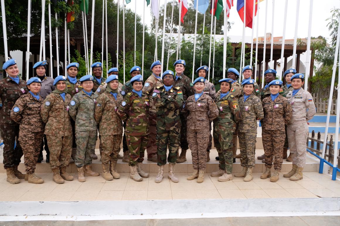 Women in military uniform stand in a row in front of flagpoles